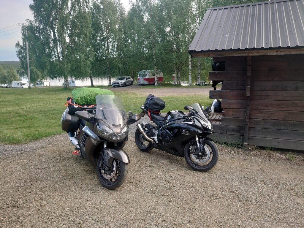 Two motorcycles (Kawasaki and Suzuki) parked at the Rantala accommodation area, with a lake and campsite in the background.