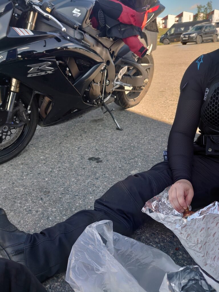 Moottoripyöräilijät pitävät taukoa Kilpisjärvellä, yksi syö eväitä maassa istuen, Suzuki GSX-R 600 vieressä parkissa.