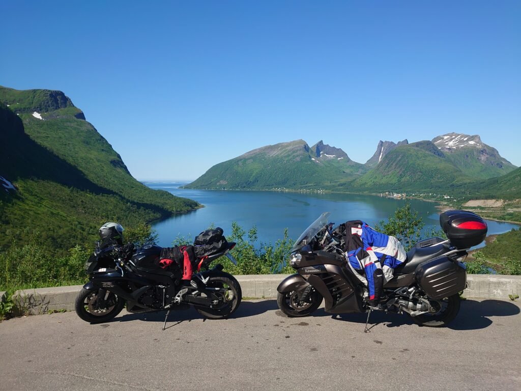 Two motorcycles parked at a scenic viewpoint, with a deep fjord and mountains of Senja in the background on a clear day.