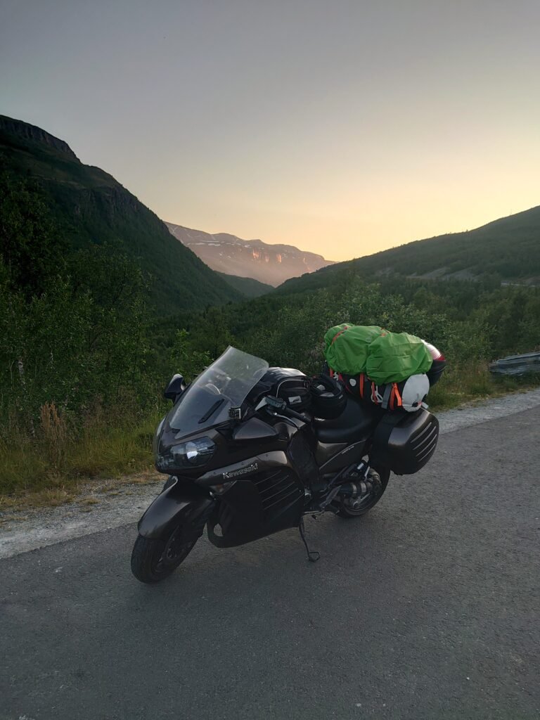 Moottoripyörä Kawasaki GTR 1400 pysäköitynä vuoristotielle iltavalossa, pyörän päällä suuri matkavarustus ja taustalla kohoavat vihreät rinteet ja iltaruskon sävyttämä vuoristo.
